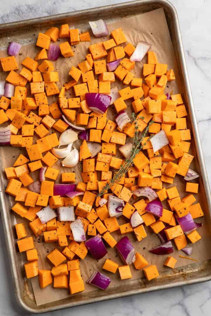 Cubed sweet potatoes, red onion, and garlic cloves seasoned with salt, pepper, cumin, cinnamon, and thyme sprigs on a sheet pan.