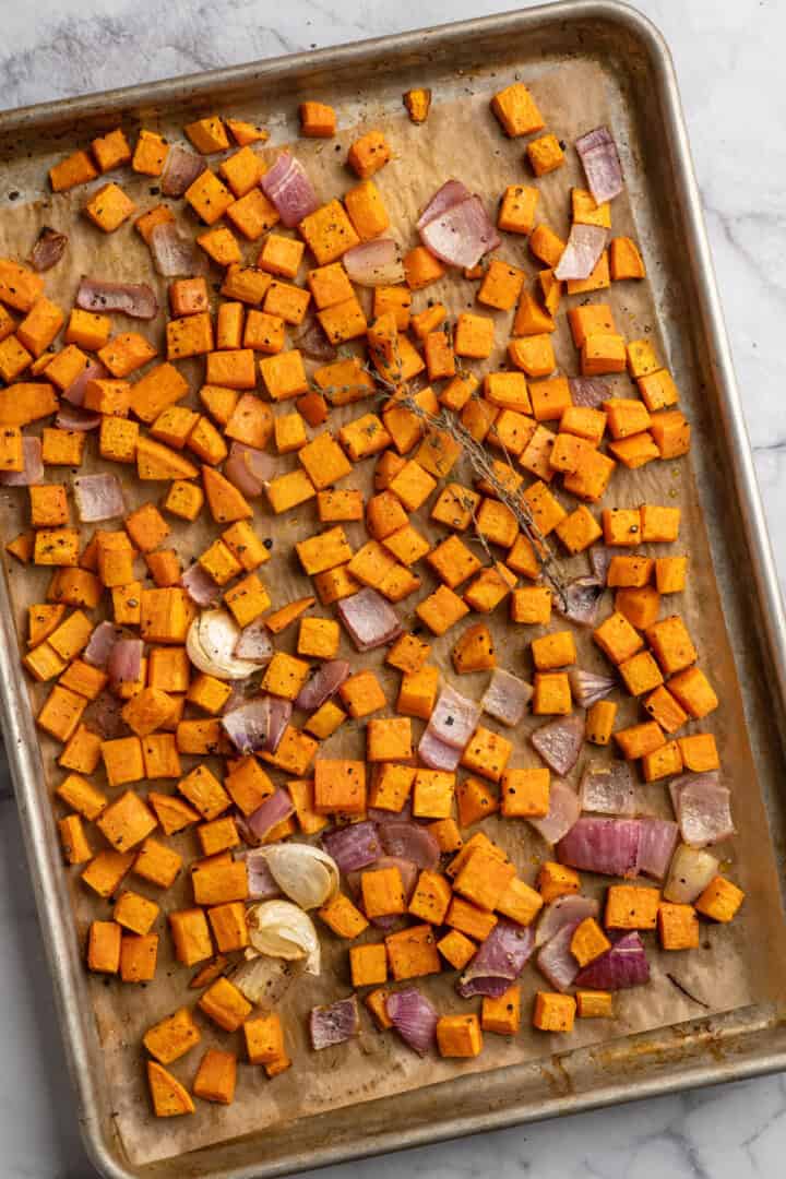 Roasted sweet potatoes, red onion, and garlic seasoned with salt, pepper, cumin, cinnamon, and thyme sprigs on a sheet pan.