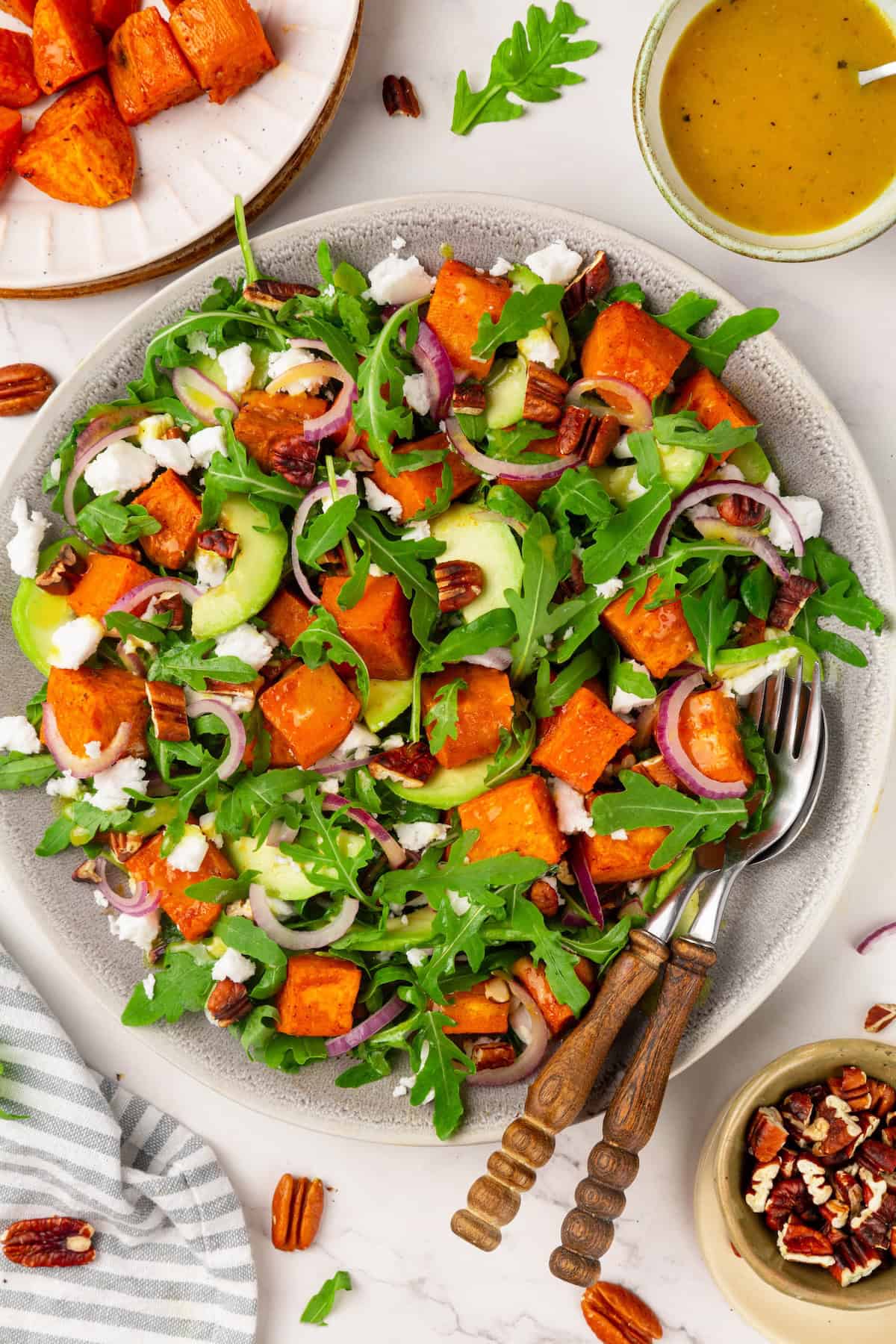 This sweet 'n savoury sweet potato salad is full of warm, tender sweet potatoes, crisp, peppery arugula, toasted pecans, and tangy vegan feta. Overhead image of sweet potato arugula salad on a serving platter with a fork and a spoon.