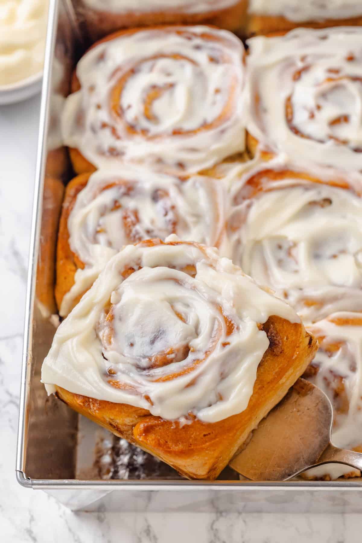 Fabulously fluffy, filled with soft, cinnamony apples, and topped with cream cheese frosting, you need these apple cinnamon rolls now. A spatula lifting a vegan apple cinnamon roll out of a baking pan.