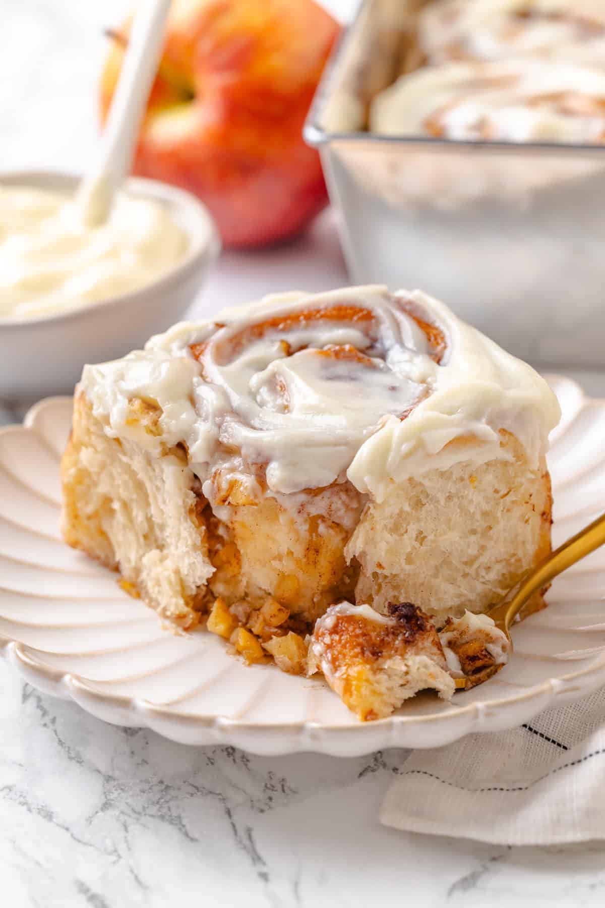 Fabulously fluffy, filled with soft, cinnamony apples, and topped with cream cheese frosting, you need these apple cinnamon rolls now. An apple cinnamon roll on a plate with a fork holding a bite of it.