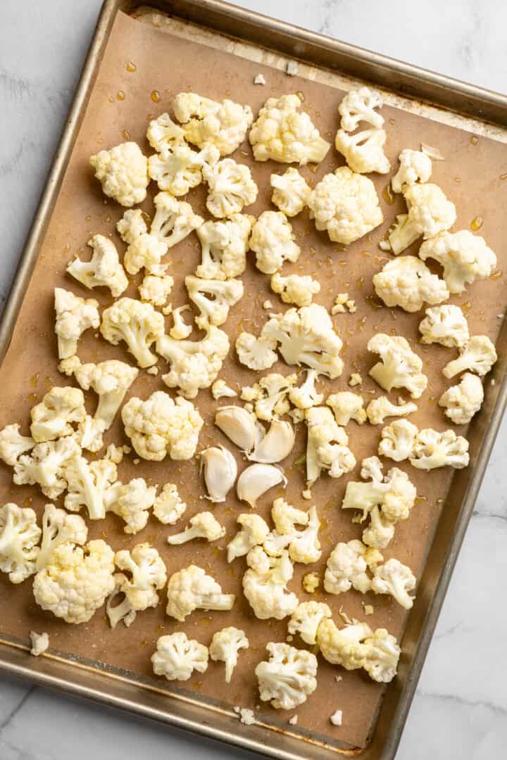 This cauliflower mash is made with roasted cauliflower and garlic for the ultimate sauvoury flavour, and coconut milk for a silky finish. Cauliflower florets and garlic cloves spread out on a baking sheet.