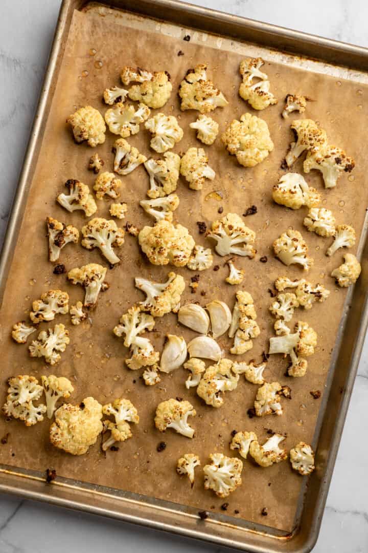 This cauliflower mash is made with roasted cauliflower and garlic for the ultimate sauvoury flavour, and coconut milk for a silky finish. Roasted cauliflower florets and garlic cloves spread out on a baking sheet.
